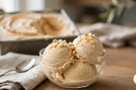 Three scoops of Amarula ice cream with a caramel drizzle and chopped macadamia nuts in a glass bowl, with the caramel-swirled loaf pan behind