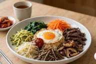 A wide white ceramic bowl of bibimbap on a light oak table, rice topped with separate sections of seasoned spinach, julienned carrots, zucchini matchsticks, bean sprouts, fernbrake, glossy shiitake mushrooms and seared beef around a sunny-side-up egg sprinkled with sesame seeds, a spoonful of red gochujang beside the egg, with a dish of kimchi, a cup of barley tea, a steel spoon and chopsticks nearby