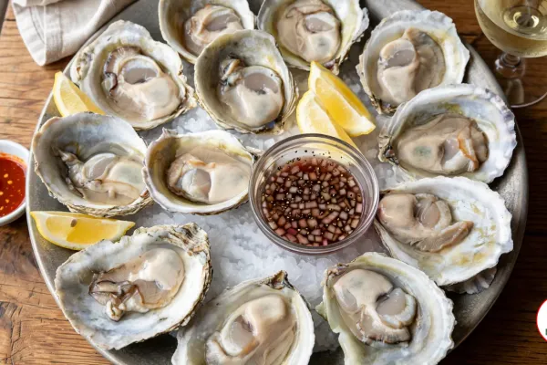 A pewter platter of a dozen raw Bluff oysters on crushed ice on a wooden table, round flat shells holding plump creamy oysters, with lemon wedges and a glass bowl of shallot mignonette in the middle, a dish of chili hot sauce and a glass of white wine beside it
