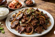 A white plate piled with Korean bulgogi, thin slices of caramelized beef with charred edges tangled with soft onion and green onion and sprinkled with sesame seeds and sliced green onion, with a bowl of steamed white rice, red leaf lettuce, a plate of napa cabbage kimchi, a dish of ssamjang, sliced garlic and sliced green chili on a rustic wooden table