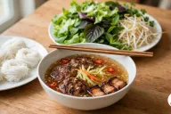 A bowl of nuoc cham with charcoal-grilled pork patties, pork belly and pickled carrot and papaya, chopsticks across the rim, beside plates of rice vermicelli and fresh herbs on a wooden table
