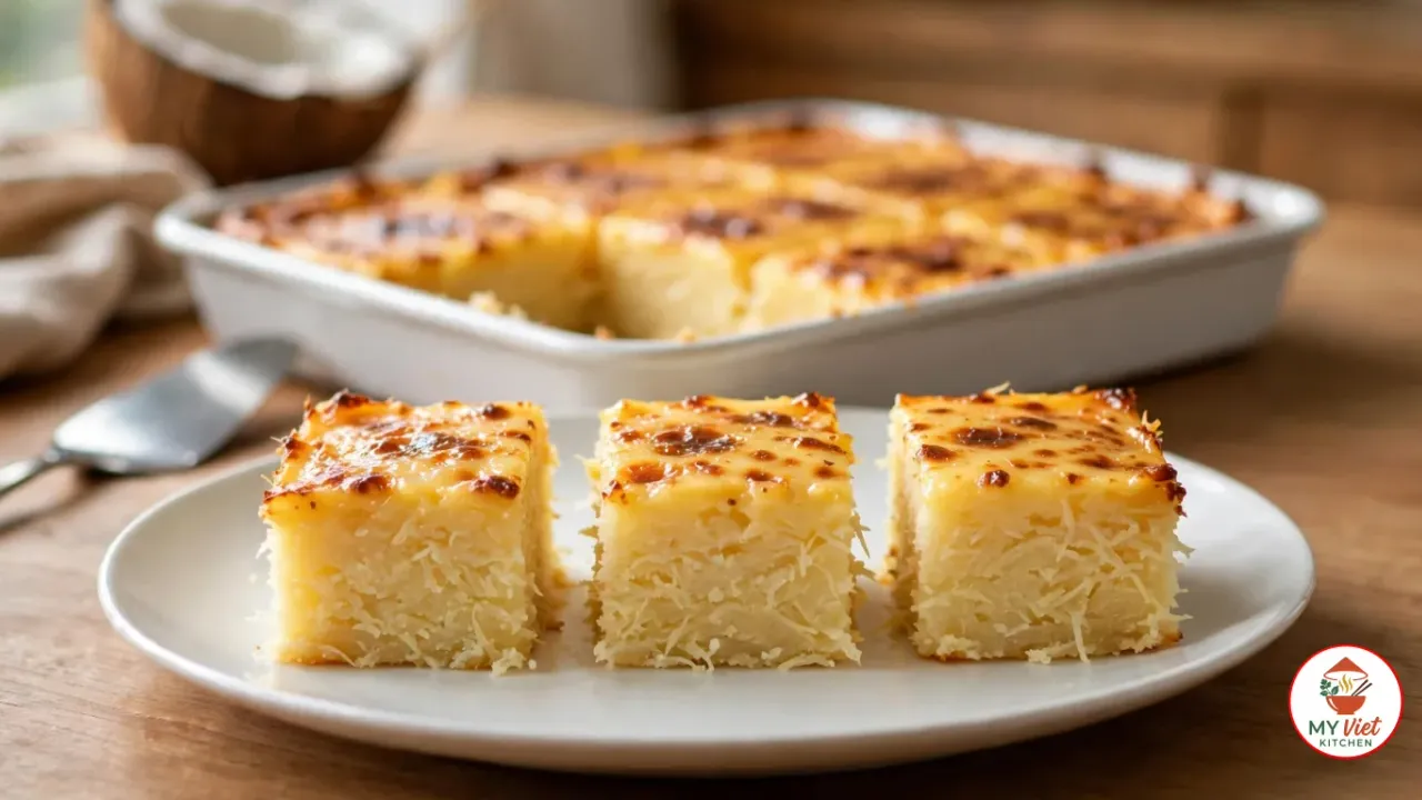 Three squares of cassava cake with a golden, brown-spotted custard top on a white plate, in front of the rest of the cake in a white rectangular baking dish, with a cake server, half a coconut and a linen napkin on a wooden table