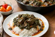A white plate of long-grain rice topped with dark green cassava leaf stew, chunks of beef and flakes of smoked fish, with orange-red palm oil pooling around it, in front of a black cast-iron pot of the stew and a small bowl of habanero peppers on a wooden table