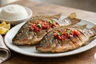 Two whole pan-fried chambo with crisp, deeply scored golden skin on a white oval platter, topped with tomato and onion relish and cilantro, beside a bowl of white nsima, lemon wedges and a linen napkin on a wooden table
