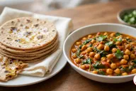 A stack of whole wheat chapatis with brown spots and a sheen of ghee on a cream cloth on a white plate, one torn in half, beside a white bowl of chickpea and spinach curry in an orange-red sauce topped with cilantro, on a wooden table