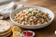 A wide white bowl of chicken kelaguen, chopped grilled chicken with charred edges flecked with grated coconut, green onion, diced white onion and sliced red chili, beside a stack of flour titiyas, two lemon halves and a small dish of red bird's eye chilies on a wooden table