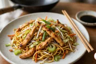 A speckled off-white stoneware plate of chicken chow mein, thin egg noodles tossed with seared strips of chicken thigh, shredded cabbage, sliced celery, carrot matchsticks, bean sprouts and green onion, with wooden chopsticks, a dish of soy sauce and a black wok behind on a wooden table