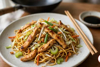 A speckled off-white stoneware plate of chicken chow mein, thin egg noodles tossed with seared strips of chicken thigh, shredded cabbage, sliced celery, carrot matchsticks, bean sprouts and green onion, with wooden chopsticks, a dish of soy sauce and a black wok behind on a wooden table