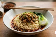 A wide white speckled bowl of dan dan noodles, the pale wheat noodles sitting in a shallow pool of red chili oil and topped with crisp minced pork and dark ya cai, crushed peanuts, sliced green onion and half a baby bok choy, with wooden chopsticks across the rim on a wooden table
