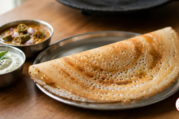A crisp golden-brown dosa folded into a half-moon on a round stainless-steel plate, its lacy surface full of tiny holes, beside small steel bowls of white coconut chutney topped with curry leaves and mustard seeds and of sambar with okra and shallots, a black cast-iron tawa behind on a wooden table