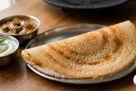 A crisp golden-brown dosa folded into a half-moon on a round stainless-steel plate, its lacy surface full of tiny holes, beside small steel bowls of white coconut chutney topped with curry leaves and mustard seeds and of sambar with okra and shallots, a black cast-iron tawa behind on a wooden table