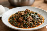 A wide white speckled ceramic bowl of efo riro, chopped dark green spinach in a coarse orange-red palm oil and pepper sauce with cubes of beef, flaky chunks of smoked fish and pale grey pieces of stockfish, beside a smooth ball of pounded yam on a small plate and a steel pot on a wooden table