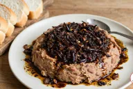 A mound of pale tan mashed beans on an off-white plate, topped with a very dark, almost black agoyin sauce of fried onions and red pepper flakes, its dark oil pooling on the plate, with a spoon on the rim and slices of soft white agege bread on a wooden board
