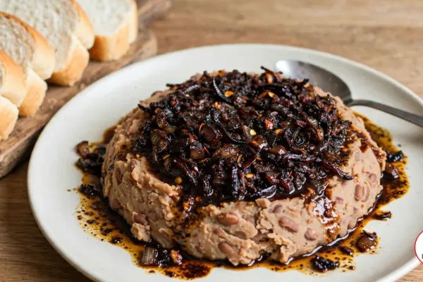 A mound of pale tan mashed beans on an off-white plate, topped with a very dark, almost black agoyin sauce of fried onions and red pepper flakes, its dark oil pooling on the plate, with a spoon on the rim and slices of soft white agege bread on a wooden board