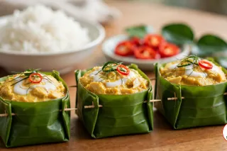 Three folded banana-leaf cups of yellow fish amok pinned with wooden toothpicks, each topped with coconut cream, fine threads of kaffir lime leaf and a ring of red chilli, with a bowl of jasmine rice and a dish of sliced red chilli behind them on a wooden table