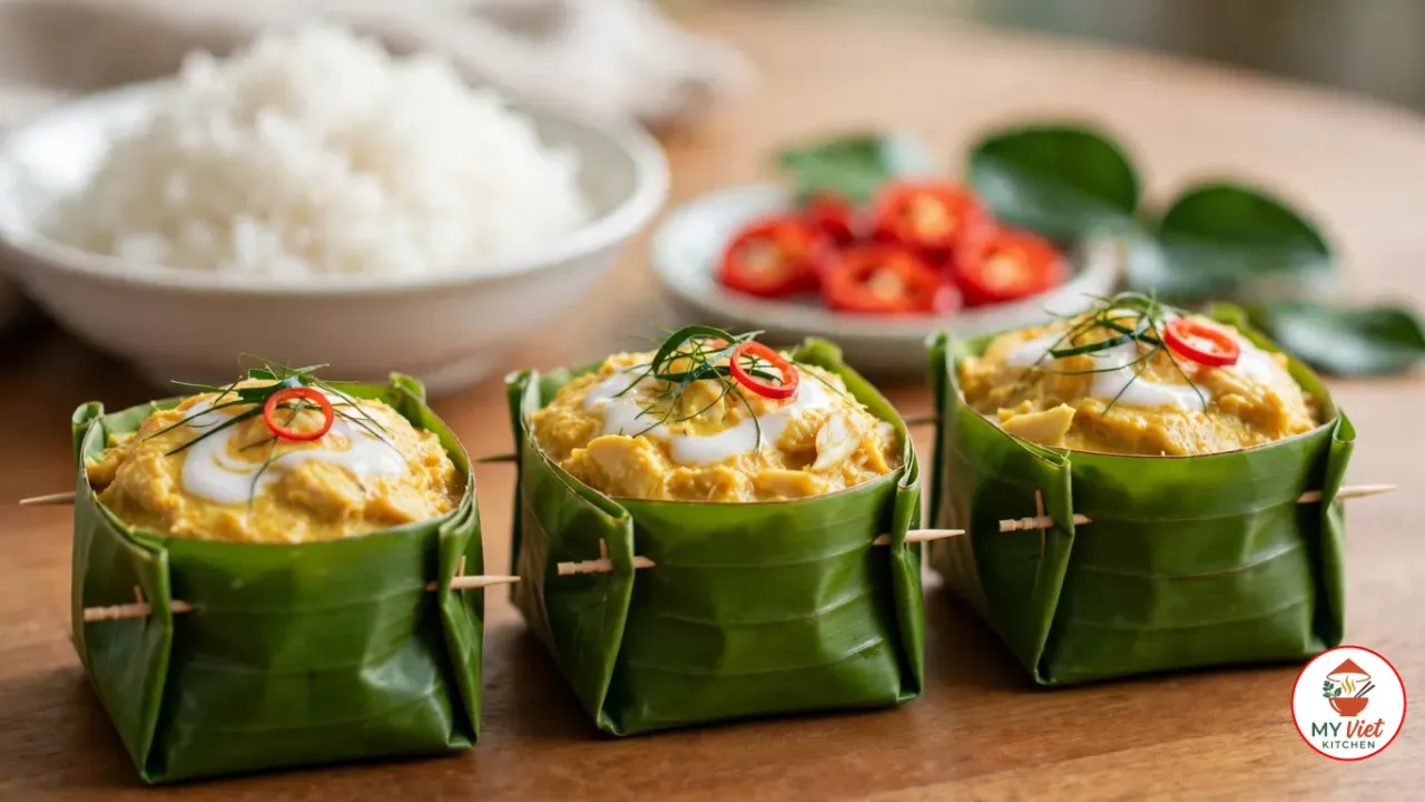Three folded banana-leaf cups of yellow fish amok pinned with wooden toothpicks, each topped with coconut cream, fine threads of kaffir lime leaf and a ring of red chilli, with a bowl of jasmine rice and a dish of sliced red chilli behind them on a wooden table