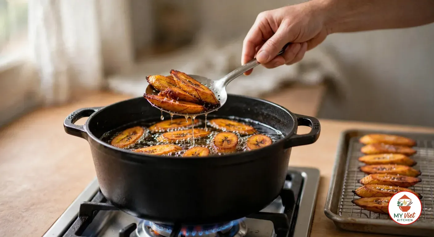 Lifting golden fried plantain slices out of the hot oil with a slotted spoon, with more alloco draining on a wire rack