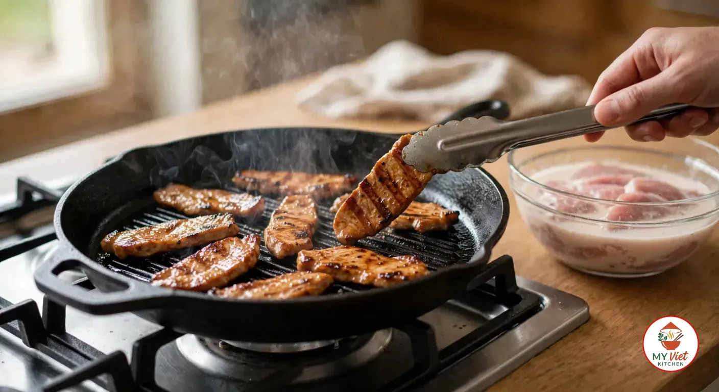 A hand turning a slice of marinated pork with metal tongs in a smoking black cast-iron grill pan on a gas stove, the slices striped with dark grill marks, beside a glass bowl of raw pork in coconut-milk marinade