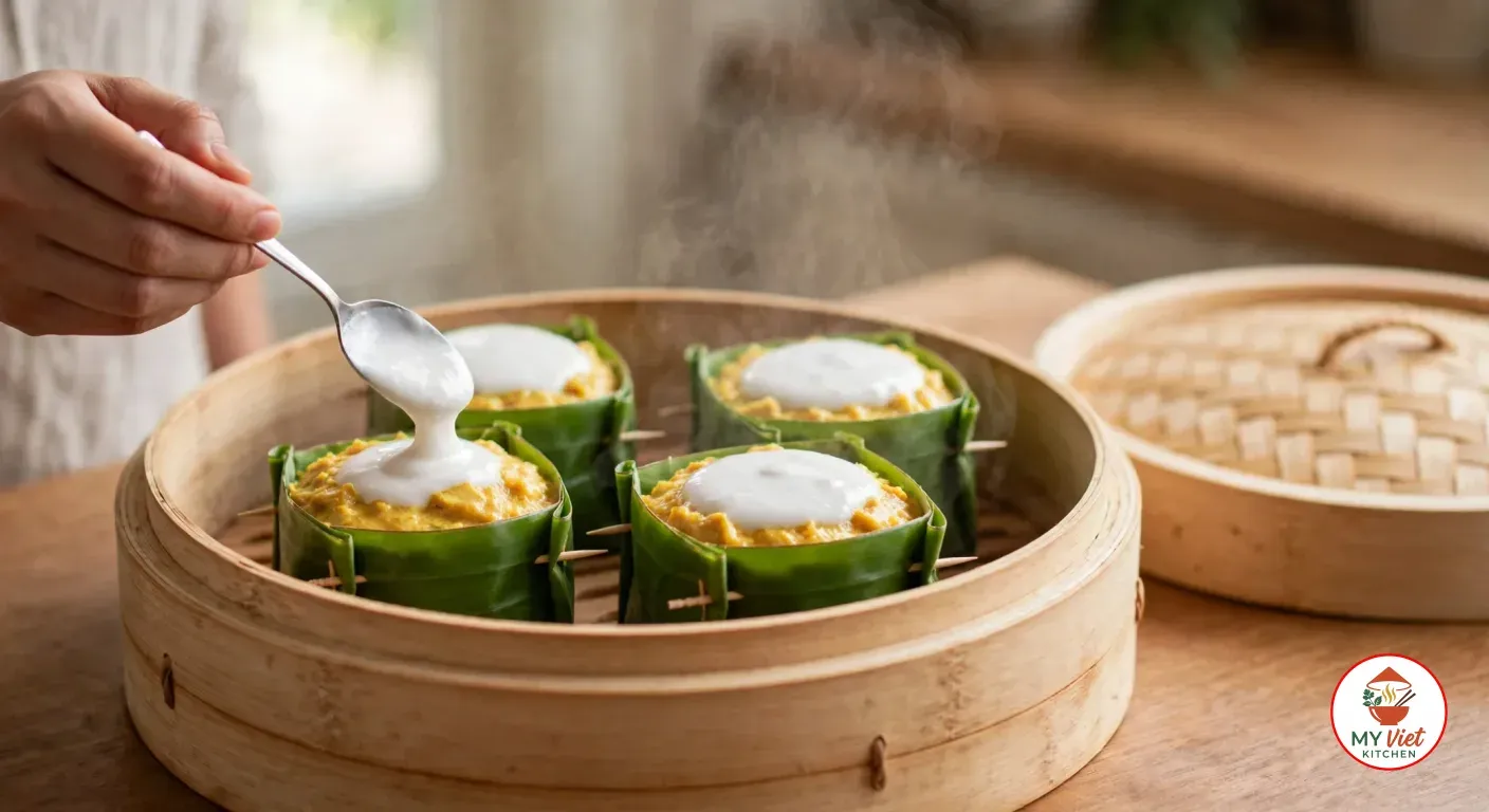 A hand spooning thick white coconut cream onto set yellow fish amok in one of four banana-leaf cups inside an open bamboo steamer, with steam rising and the woven bamboo lid beside it