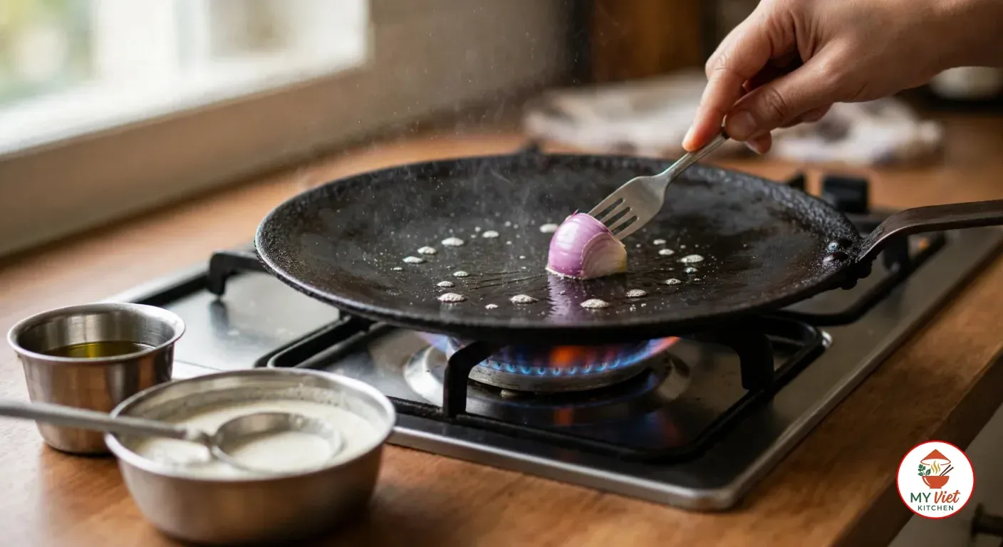A hand rubbing half a red onion on a fork over a hot black cast-iron tawa on a gas burner, water drops sizzling on the surface, with small steel bowls of oil and off-white batter on the wooden counter