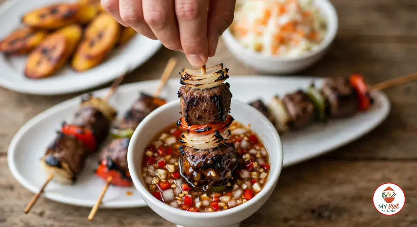 Dipping a grilled goat brochette into a small white bowl of pilipili of chopped red chili, garlic and onion in lemon juice, with more brochettes on a white oval platter, a plate of grilled ripe plantain slices and a bowl of shredded cabbage and carrot salad behind it