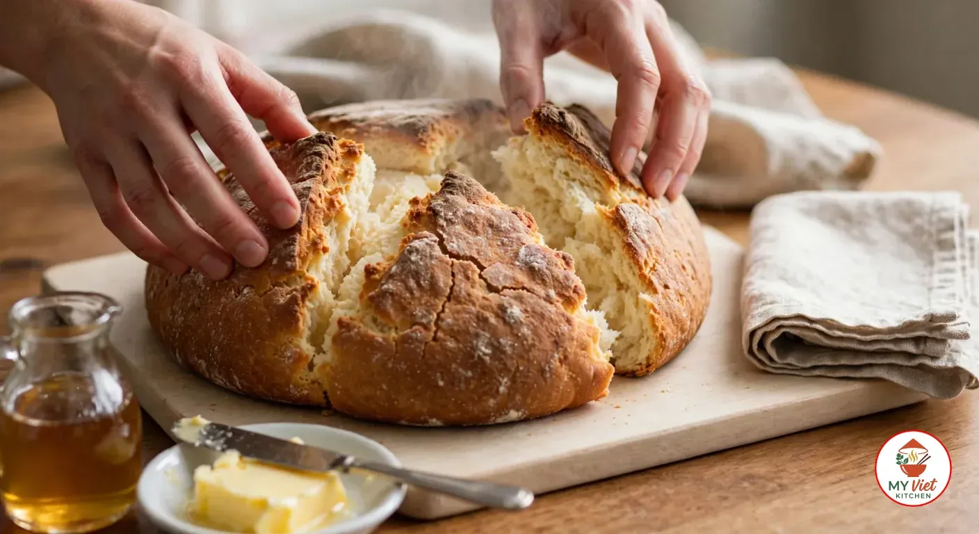 Two hands pulling a golden damper apart along its scored cross on a pale wooden board, showing the soft white crumb, with a folded linen tea towel, a dish of butter with a knife and a small glass jug of golden syrup